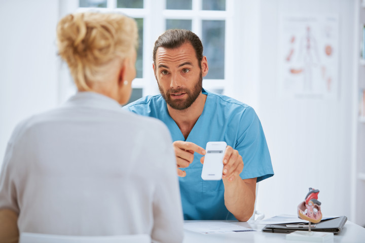 Ein medizinischer Fachangestellter klärt eine Patientin über das Implantatmonitoring auf und hält dabei den Transmitter in der Hand.